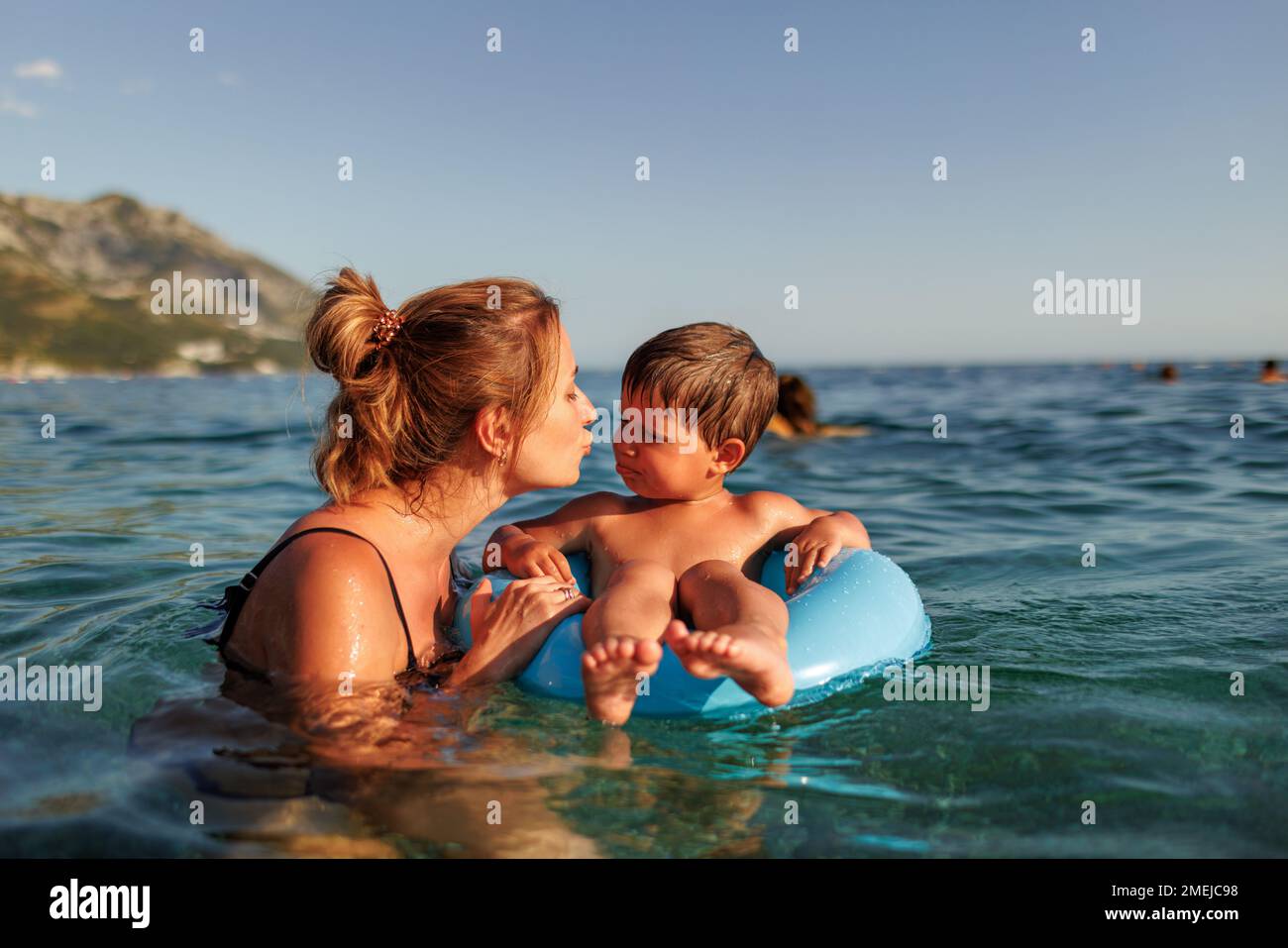 Happy young mother with blond hair plays with her little cheerful toddler son and rides him on a bright blue inflatable ring in the deep blue shiny Ad Stock Photo
