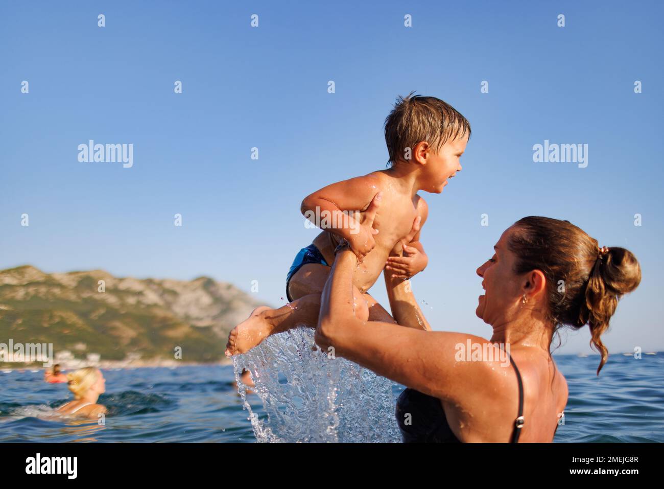 A happy mother in a bathing suit holds her little son in her arms and throws him over her head in the warm blue Adriatic Sea in the evening sunlight Stock Photo