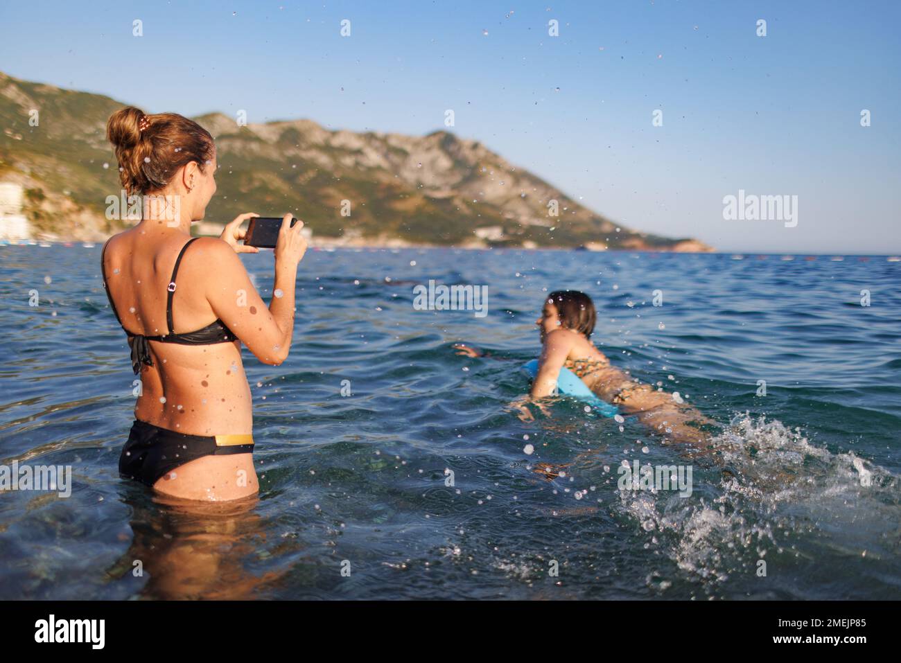 A slender caring mother takes photos on the phone en memory of her cheerful funny fooling daughter with a blue bright inflatable ring, in the summer A Stock Photo