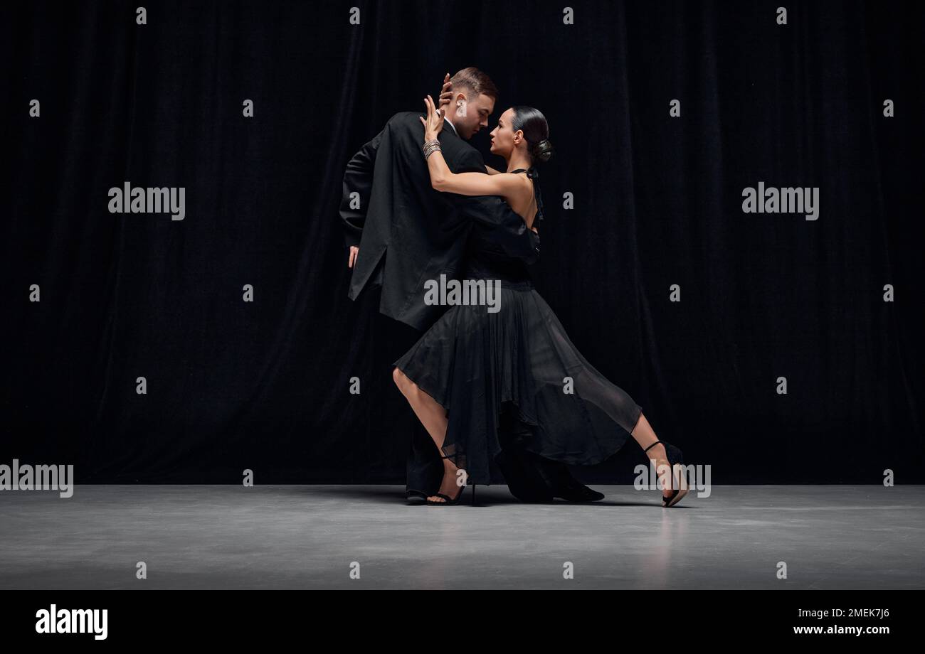 Passionate dance style. Man and woman, professional tango dancers performing in black stage costumes over black background. Stock Photo