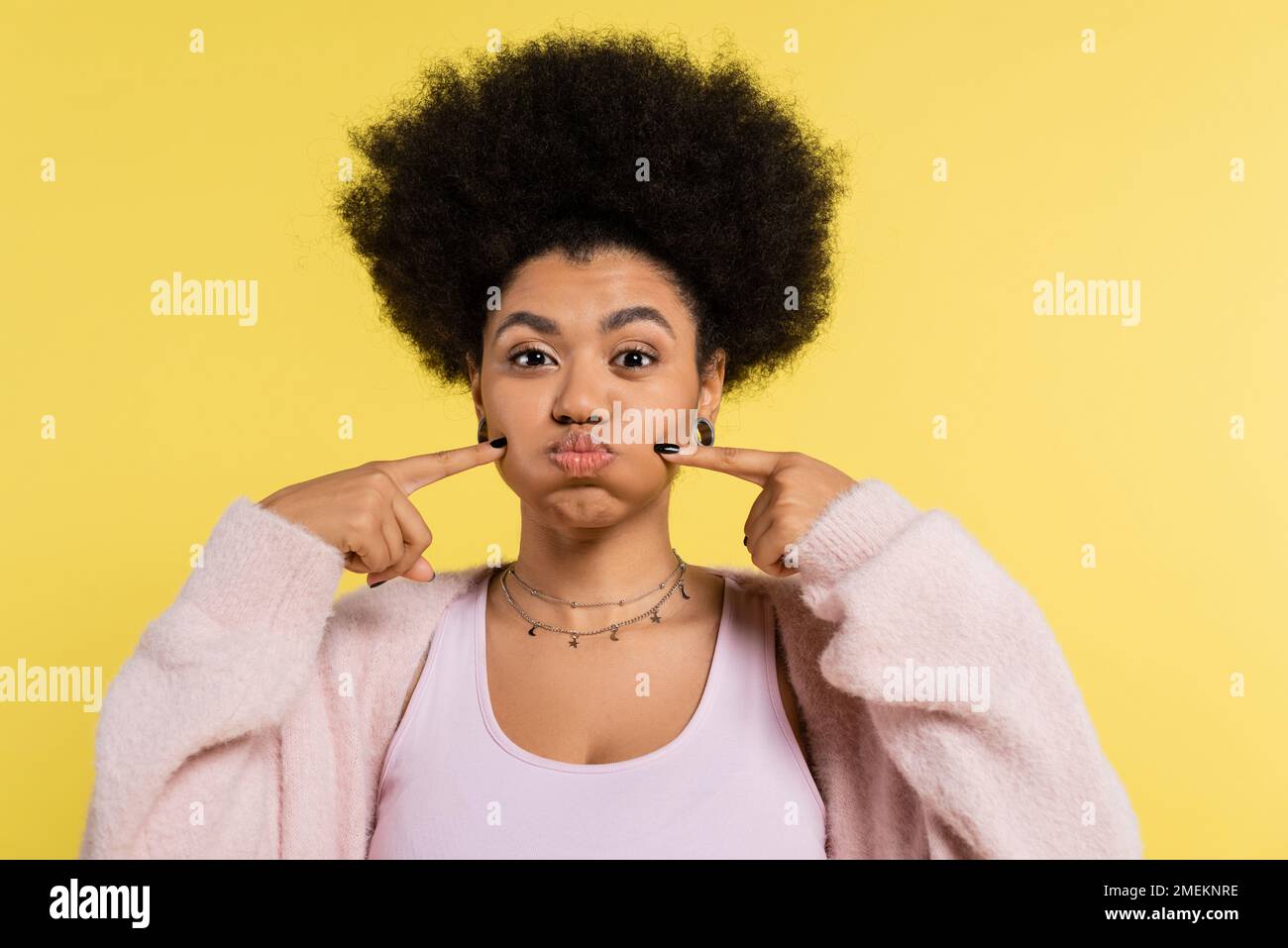 funny african american woman in silver necklaces touching puffed cheeks isolated on yellow Stock Photo
