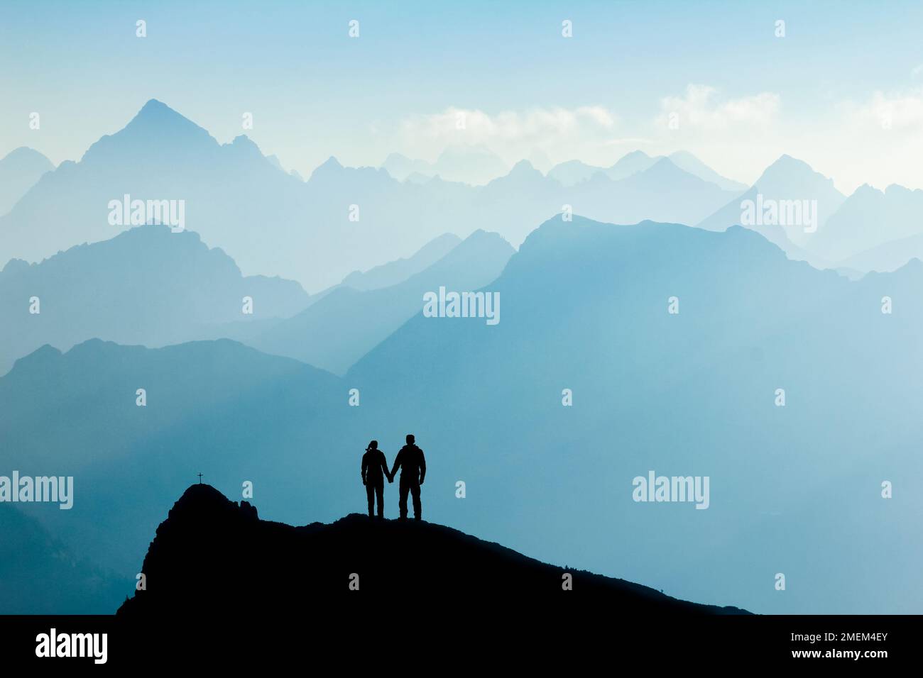 Silhouette Couple of man and woman reaching mountain top enjoying freedom and looking towards blue mountain silhouettes and sunrise. Alps, Allgaeu Stock Photo