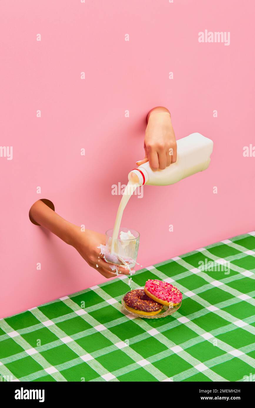 Food pop art photography. Female hands sticking out pink paper, pouring, spilling milk into cup. Yummy donuts on green tablecloth Stock Photo
