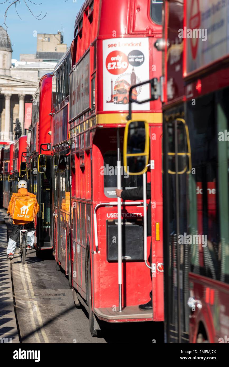 Just Eat delivery rider squeezing down the side of a queue of red London buses in a bus lane in Whitehall, Westminster, London, UK. Cyclist on inside Stock Photo