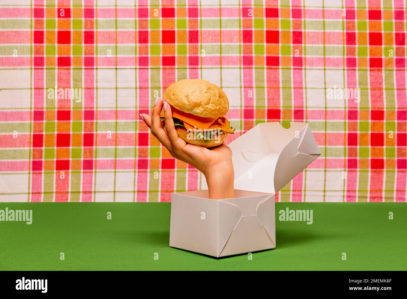 Food pop art photography. Female hand sticking out box and holding burger on green tablecloth. Junk food aesthetics Stock Photo