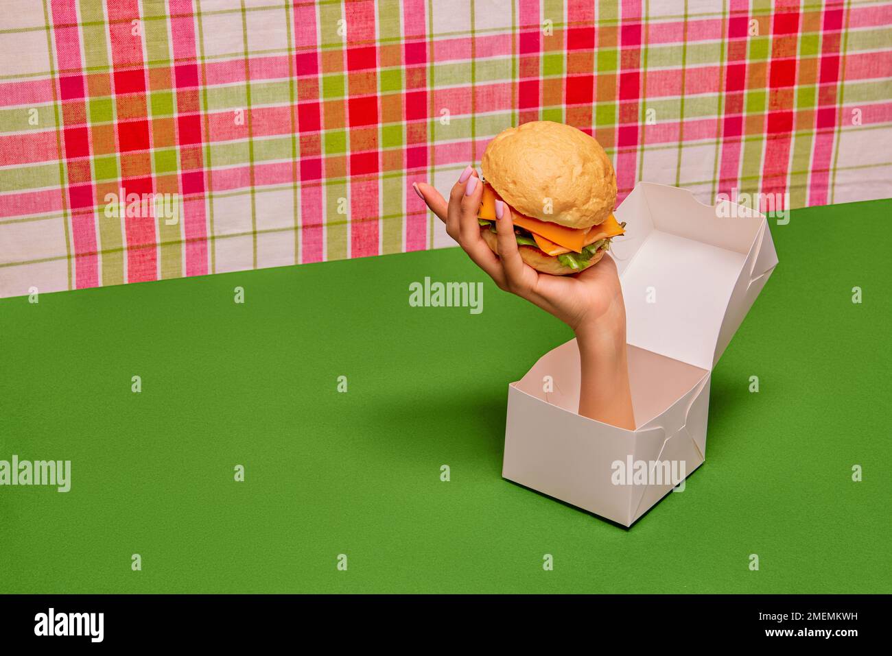 Food pop art photography. Female hand sticking out box and holding burger on green tablecloth. Delicious american food Stock Photo
