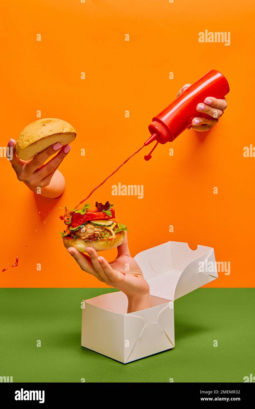 Food pop art photography. Female hand sticking out orange paper and pouring ketchup on burger on hand sticking out food box Stock Photo