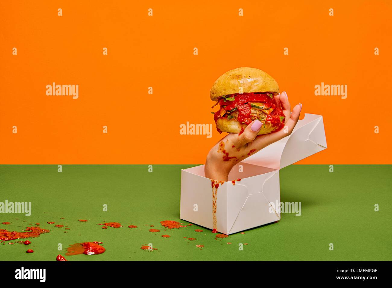 Food pop art photography. Female hand sticking out food box with burger on green tablecloth and orange background Stock Photo