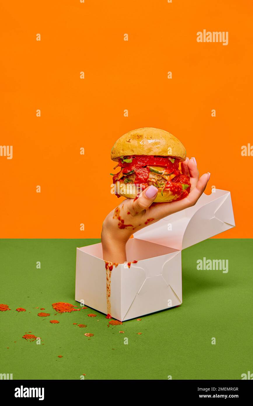 Food pop art photography. Female hand sticking out food box with delicious burger on green tablecloth and orange background Stock Photo
