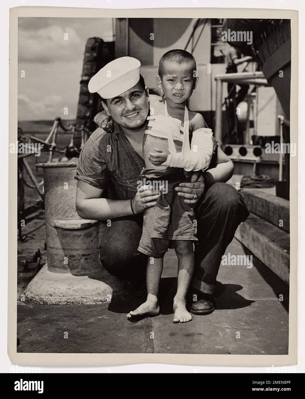Photograph of Coast Guardsman Robert Barr with a Japanese Child. De-Indoctrinated Jap. Like most Japanese children, this Nip youngster probably was indoctrinated with our enemy's philosophy before he was able to walk. But a few weeks of American kindness and medical attention accomplished a thorough job of de-indoctrination on him. The child was found injured on Saipan and brought aboard a Coast Guard-manned invasion transport. Soon he was healthy, happy devoted. He was returned to his people after the island was secured. His special pal aboard the ship was Coast Guardsman Robert Barr, with wh Stock Photo