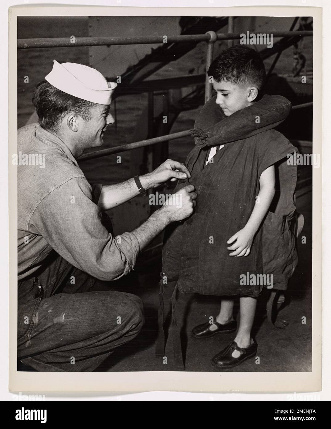 Cheer Up, Chum. Everything 'll Be Okay. A saddened little refugee boy -- his laughter lost with his home in the war-ravaged Pacific -- seems dubious about donning the life jacket aboard a Coast Guard-manned transport en route from the combat zone. To Coast Guardsman John Brady, boatswain's mate second class, the job of cheering up the youngster is tougher, in a way, than the invasion beaches of Tulagi, Guadalcanal, Rendova, and New Georgia, where Brady ran landing barges under Jap fire. The Coast Guardsman's home is in Long Island, N.Y. Stock Photo