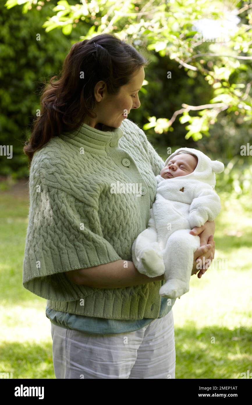 Mother standing in garden cradling sleeping baby boy (3 weeks) who is dressed in warm clothing Stock Photo