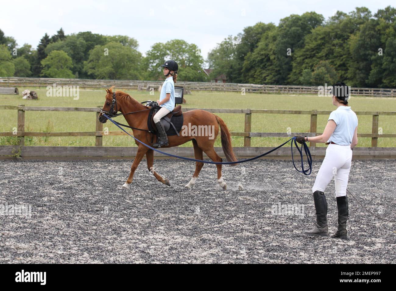 Young girl riding chestnut pony in paddock during riding lesson as instructress holds lunge rein, side view Stock Photo