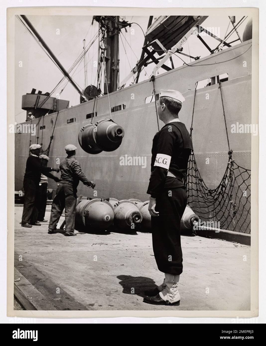 Port Security's a Coast Guard Job. Bombs Away -- First step in dropping blockbusters on the enemy is getting them to Allied airbases for the the bombardiers. Here a U.S. Coast Guard shore patrolman watches the 2,000-pound death capsules hoisted aboard ship. A large detail of Coast Guardsman constantly patrol the docks as a precaution against sabotage. Stock Photo