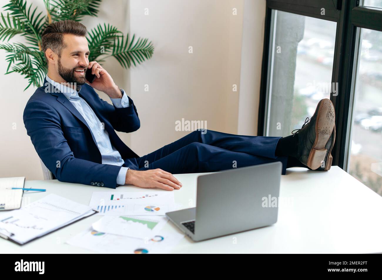 Happy elegant caucasian business man, manager, director, sits at a desk in the office, talking on the phone with a colleague or client, putting his legs on the table, making an appointment, smiling Stock Photo