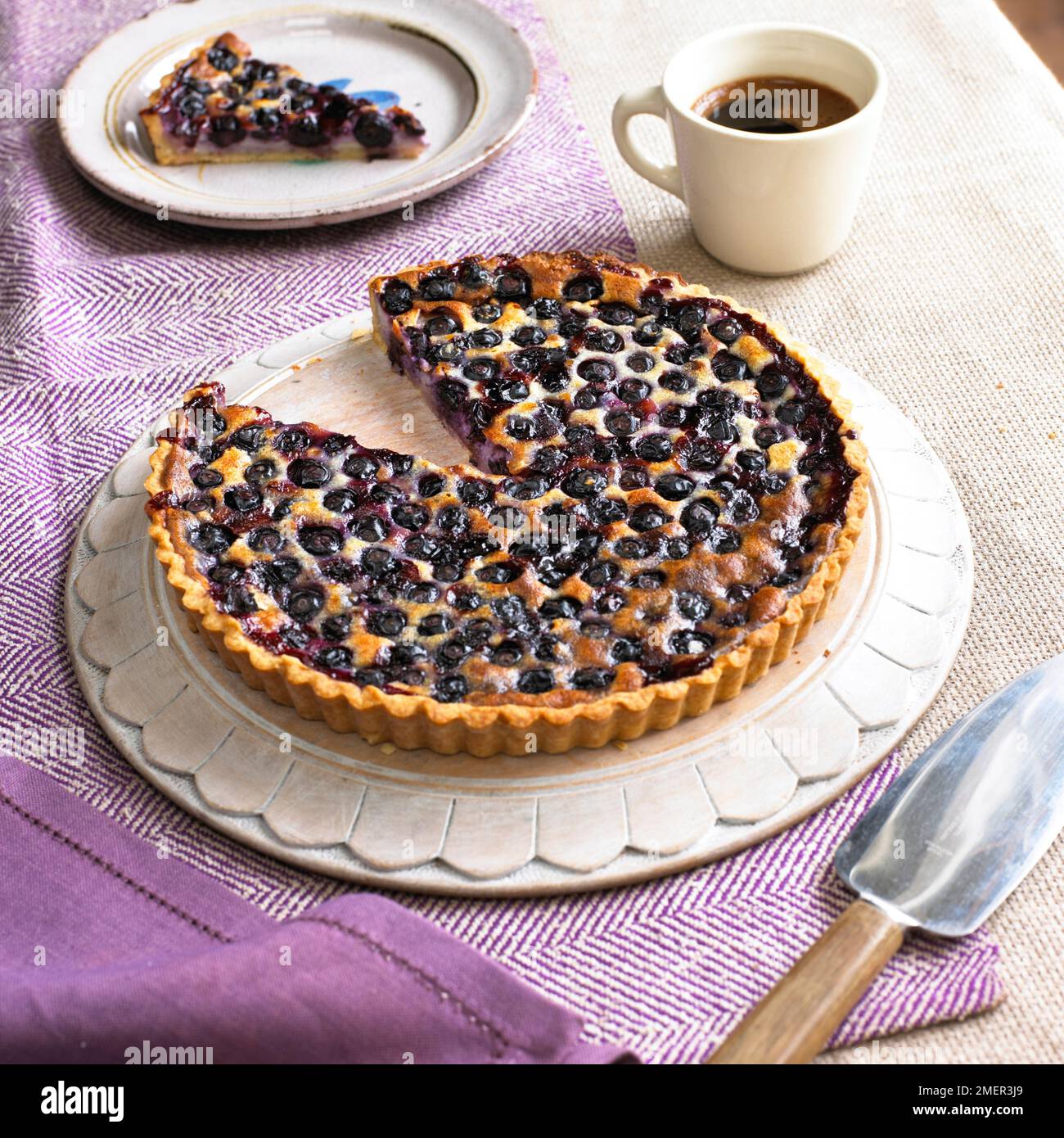Blueberry cream cheese tart with slice removed, cake server, coffee cup and slice on plate nearby Stock Photo