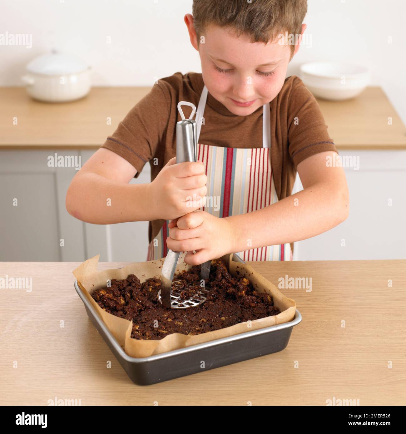 Boy using masher to press chocolate and biscuit fridge cake into cake tin, 8 years Stock Photo