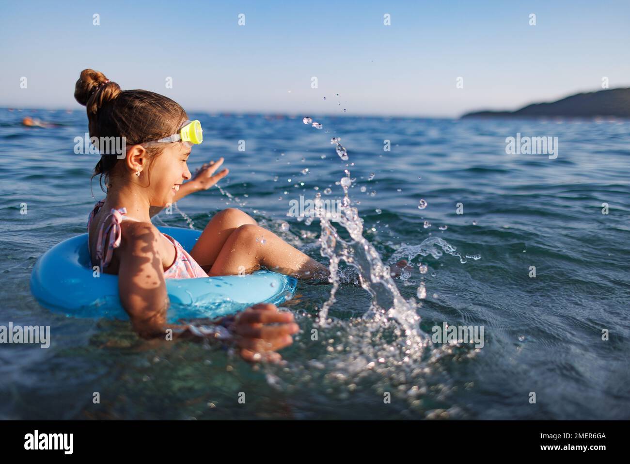 A tanned happy girl with blue swimming goggles sits in a bright blue inflatable ring and splashes with cool water while swimming, in the warm blue Adr Stock Photo