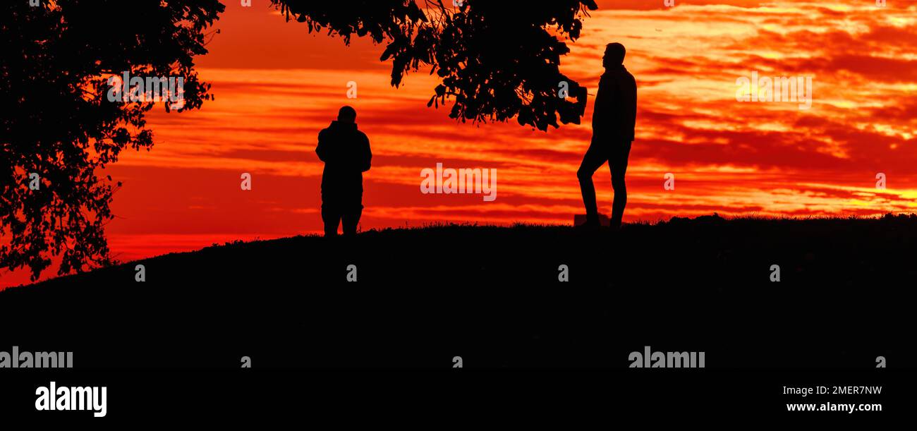 Silhouette of two men standing on the hill in sunset with vibrant orange sky in background Stock Photo