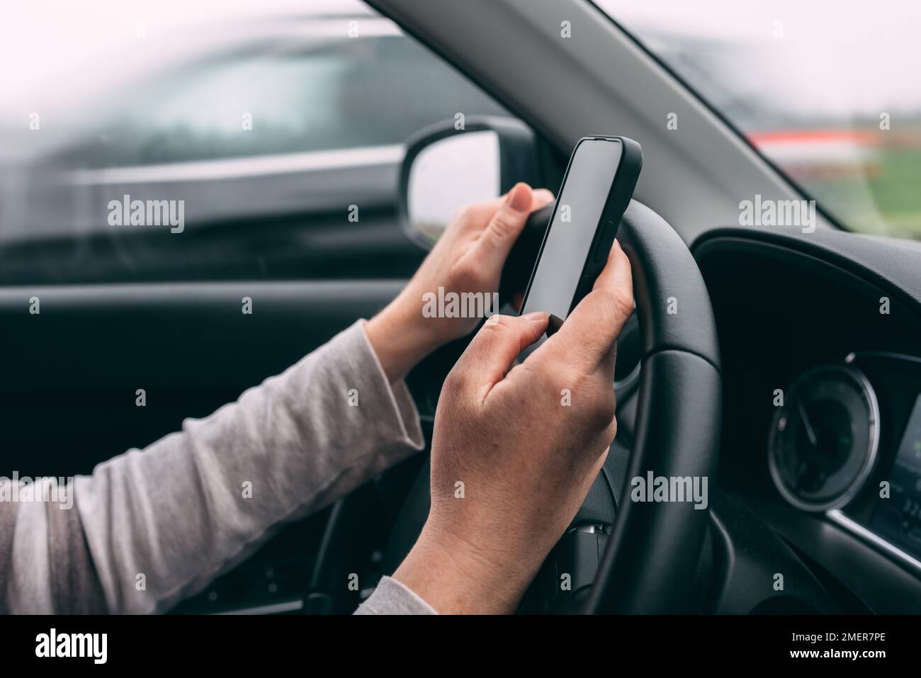 Casual adult caucasian woman typing text message on mobile smart phone while driving her car on overcast autumn day, selective focus Stock Photo