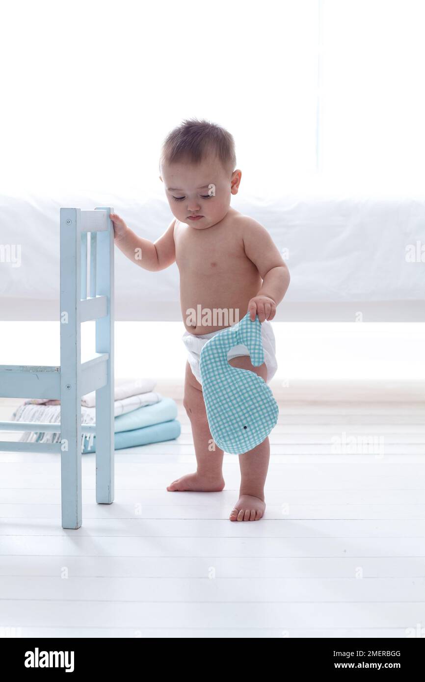 Baby boy holding cuddly whale, 11 months, standing holding on to white wooden chair Stock Photo