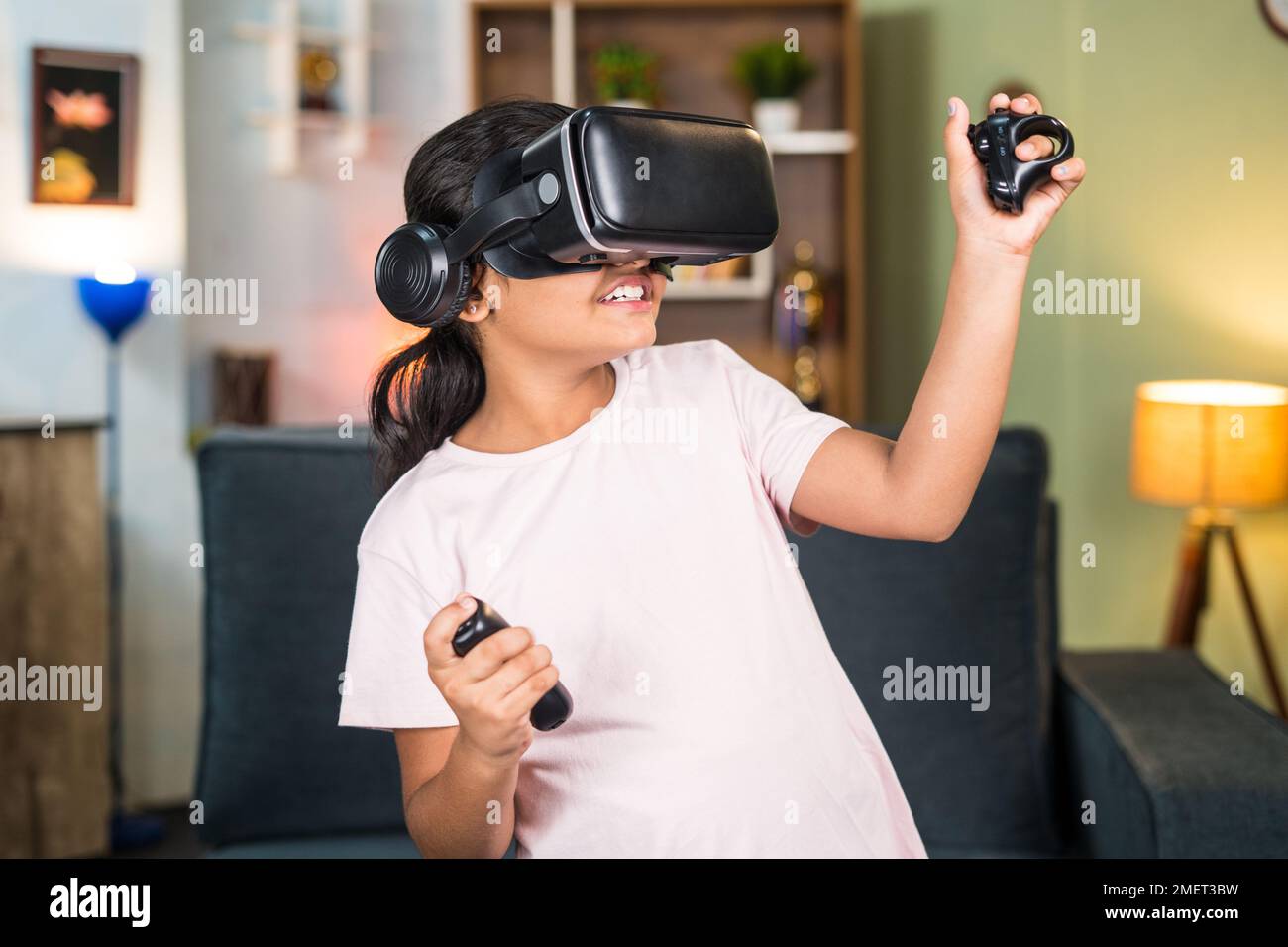 Girl child with VR or vertual reality goggles and joystick in hands playing video game on metaverse at home - concept of immersive experience Stock Photo