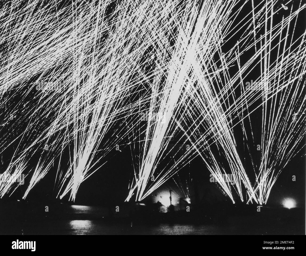 Photograph of Allied Guns Firing at Nazi Planes Off the Cherbourg Peninsula. Off Cherbourg, The Luftwaffe Attacks at Night- Allied guns weave a tapestry of flame in the night skies off the Cherbourg Peninsula, as Nazi planes appear overhead to bomb a group of invasion ships. In the foreground is the sinking hulk of an American ship, mortally wounded by a bomb hit. The glare of two bomb flashes breaks the black in the distance. This spectacular photo of battle pyrotechnics was made by a Coast Guard combat photographer under fire in this channel action. 1939 - 1967. Stock Photo