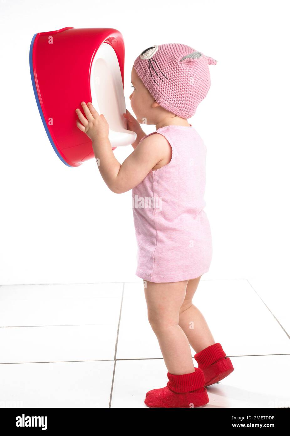 Girl standing wearing red socks, pink top and pink wool hat holding a red potty at face level, 19 months Stock Photo