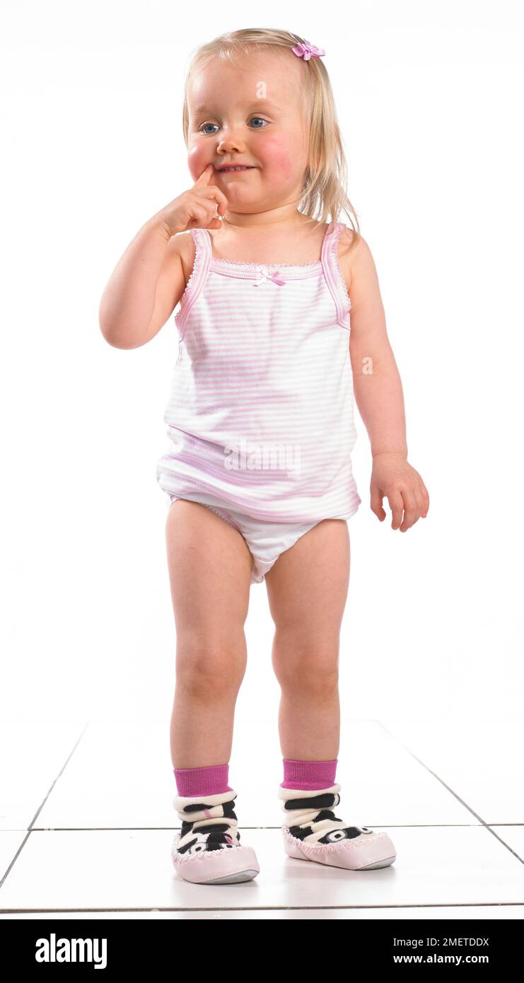 Girl wearing vest, pants and slippers, holding finger to her mouth, 20 months Stock Photo