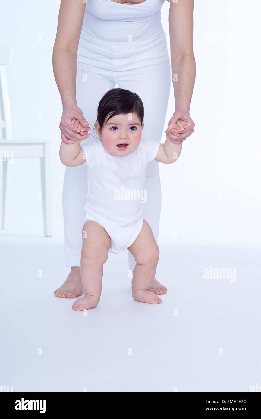Baby girl (40 weeks) standing, being supported by her mother who is standing behind her Stock