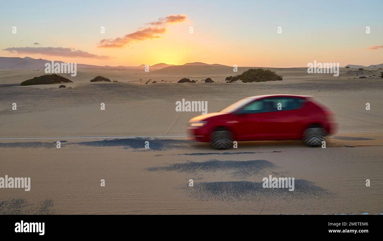 Red blurred car on sandy asphalt road, evening light, dusk, sunset, blue sky, few grey clouds, hills, northeast coast, dune area, El Jable, nature Stock Photo