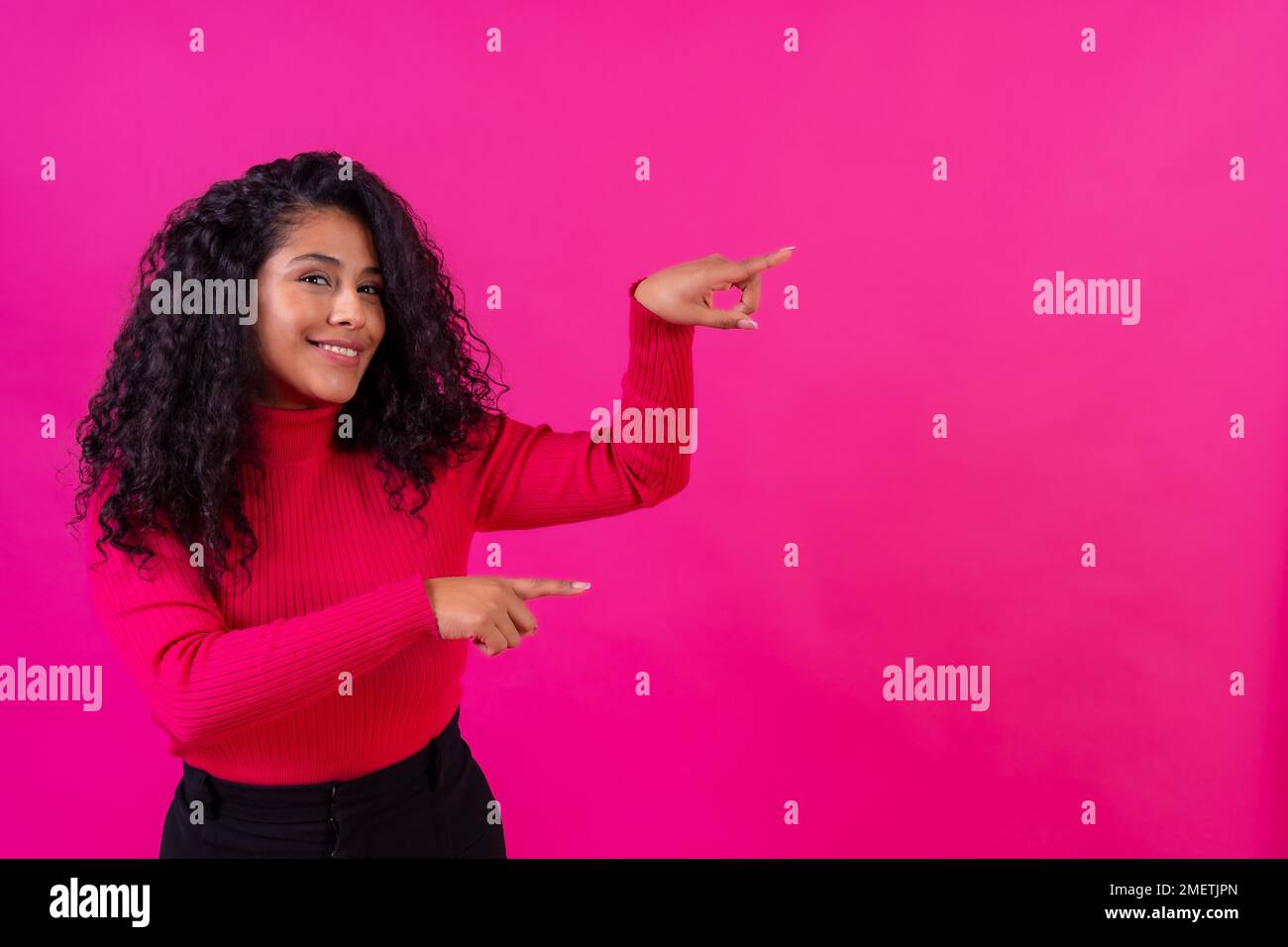 Curly-haired woman pointing to the right on a pink background, studio ...