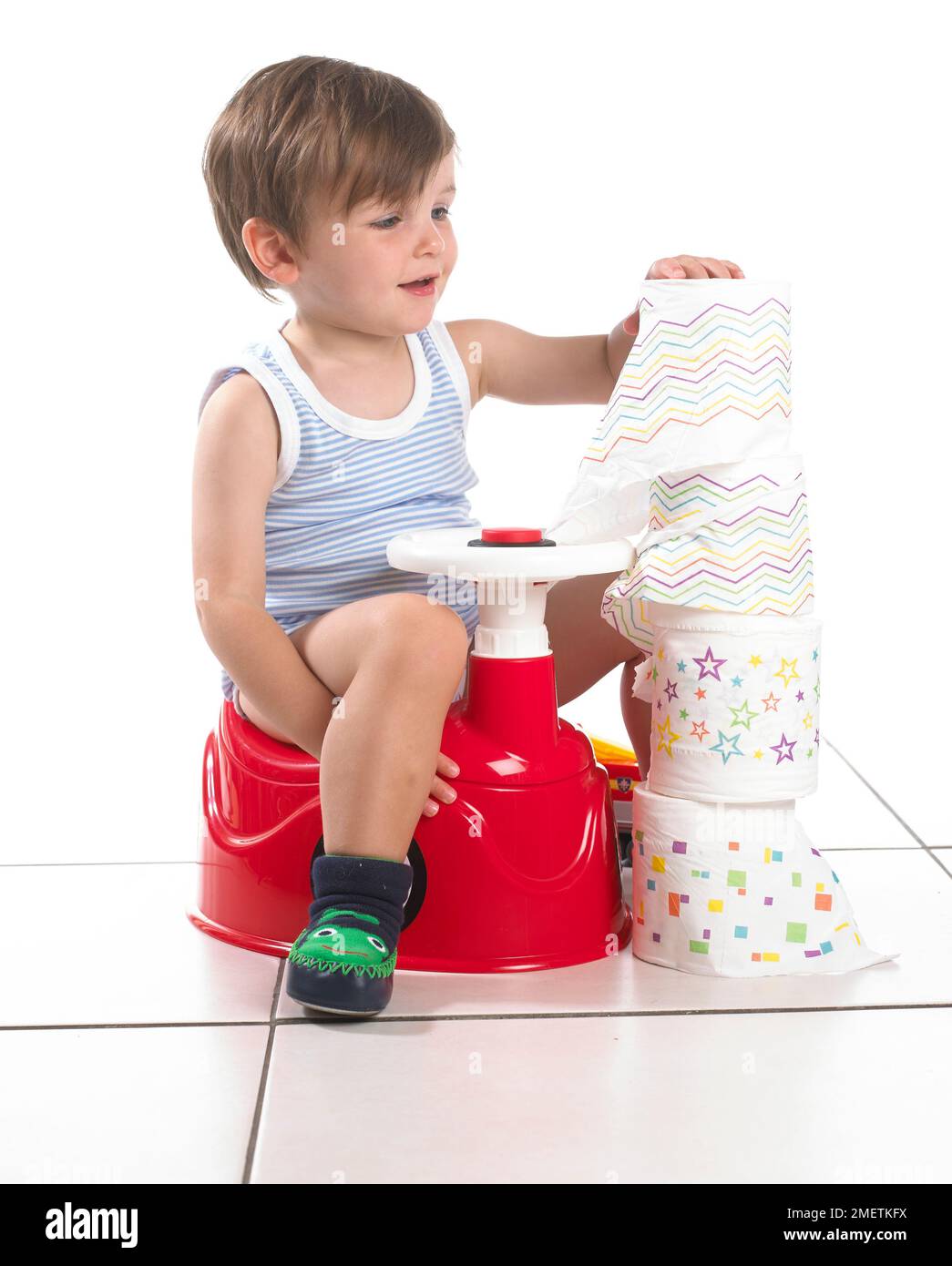 Boy sitting on a potty shaped as a car with steering wheel next to a tower of toilet rolls, 20 months Stock Photo