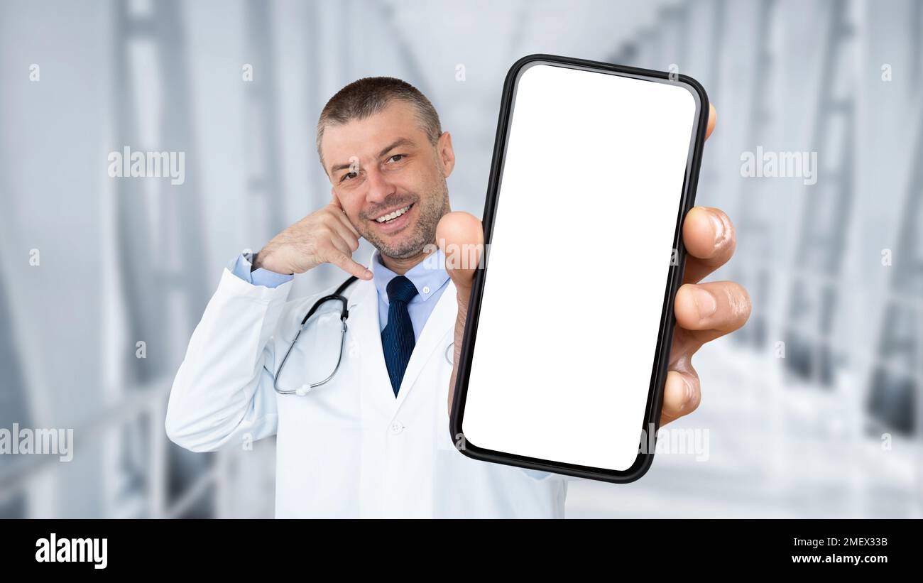 Smiling middle aged male doctor showing blank smartphone and gesturing call me Stock Photo