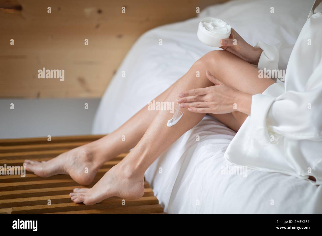 Young european woman in bathrobe with perfect legs sits on white bed with jar of cream, apply to skin Stock Photo