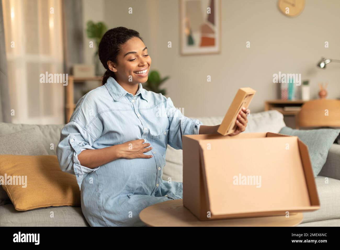 Positive pregnant black lady unpacking cardboard moving box and smiling
