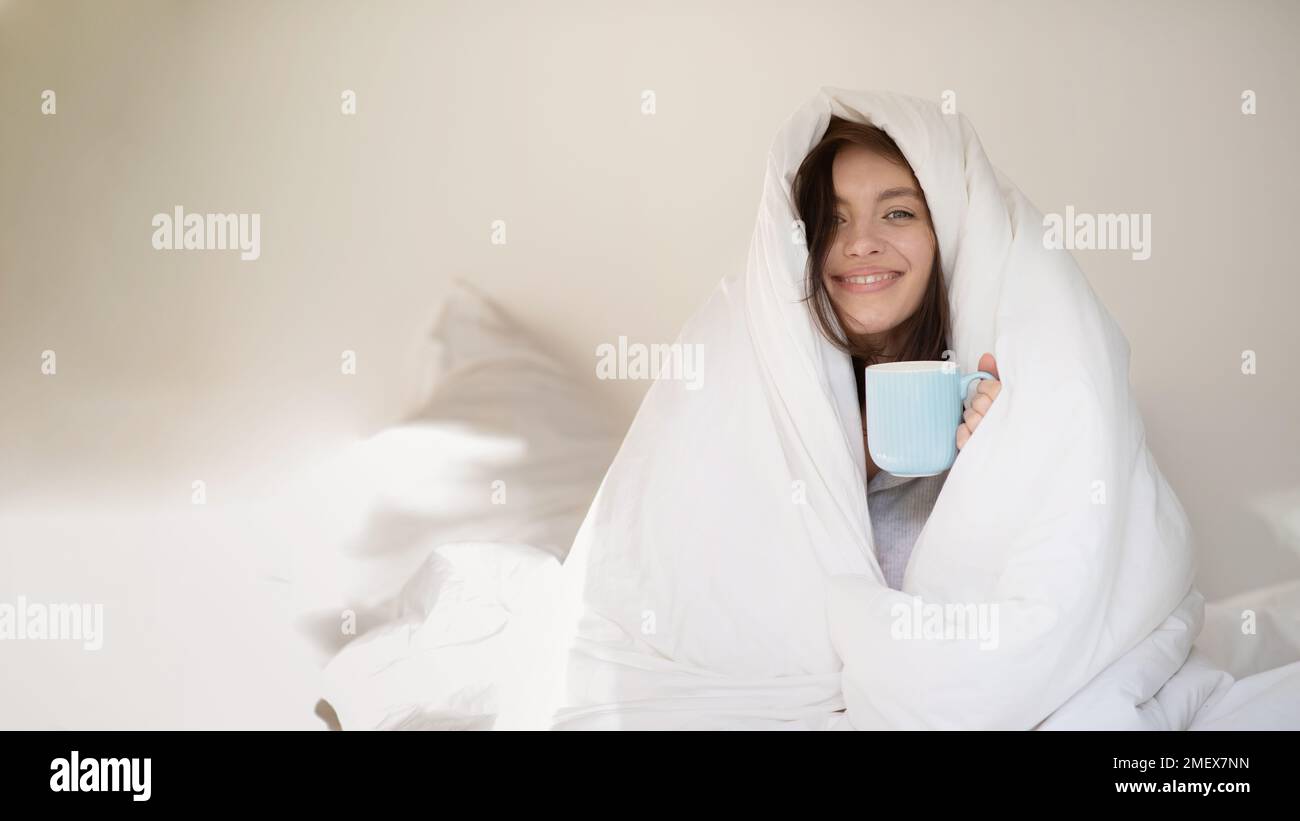 Cheerful young european woman wrapped in white blanket sits on white bed with cup of coffee Stock Photo