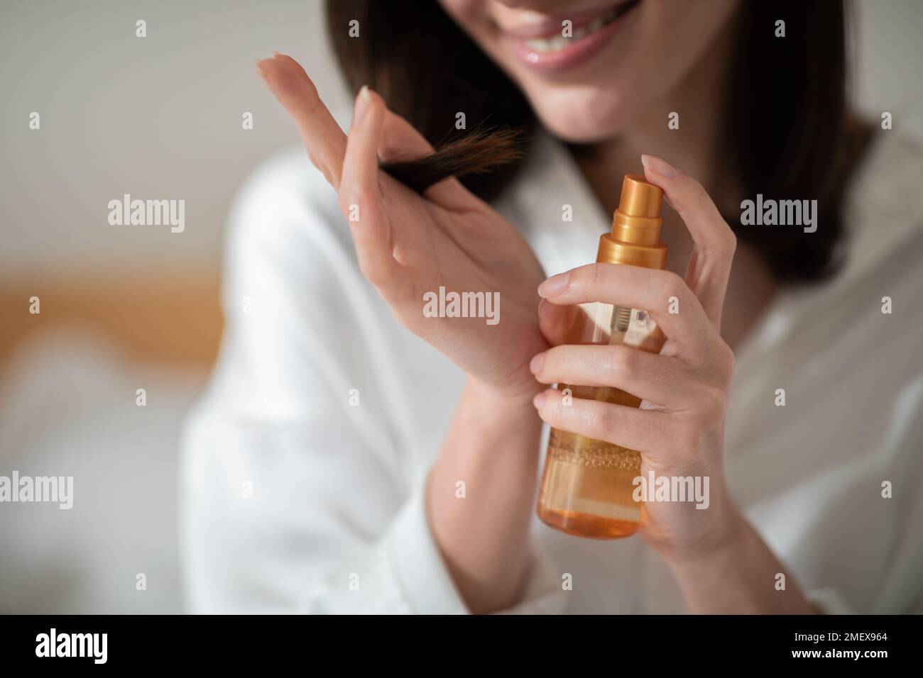 Cheerful young european woman on white bed applies spray on hair in bedroom interior, close up, cropped Stock Photo