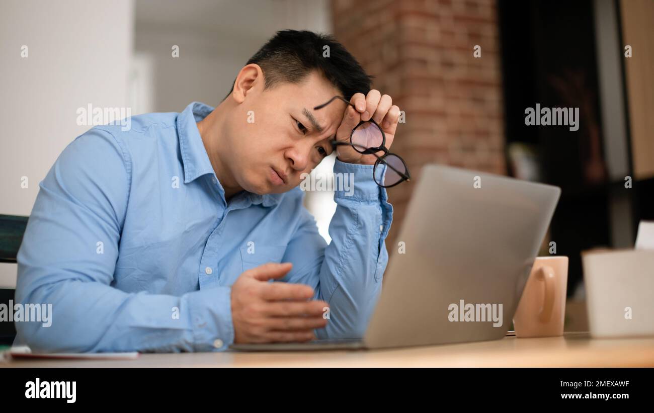 Boring tired mature asian businessman working on laptop computer, sitting at table at home office, panorama Stock Photo