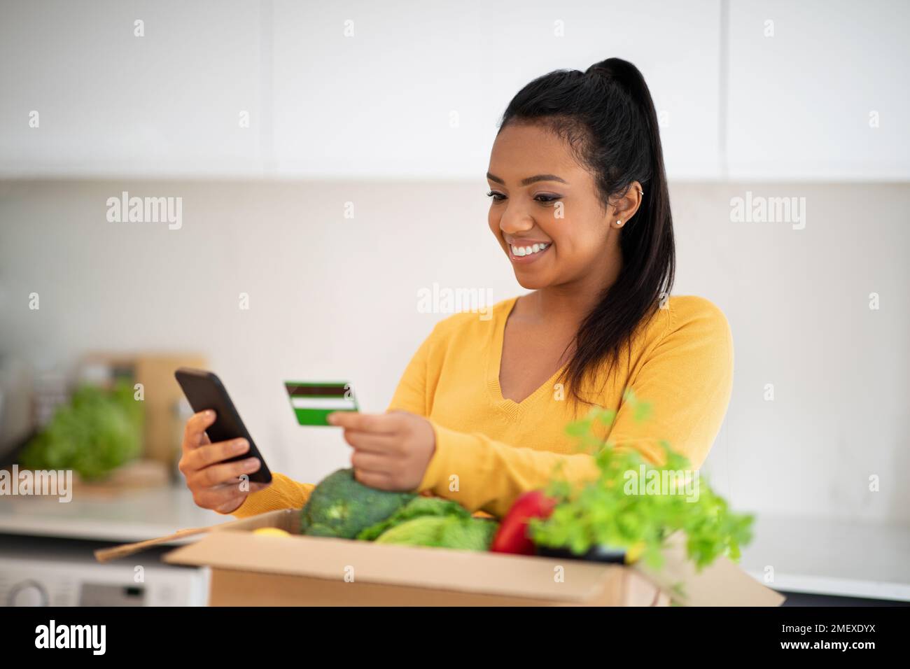 Glad millennial african american woman unpacks cardboard box with organic vegetables uses smartphone Stock Photo