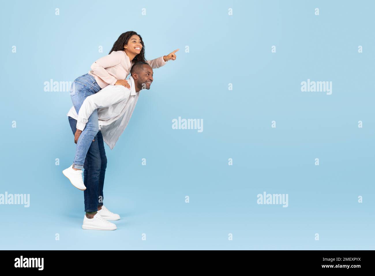 Happy black couple having fun, lady piggybacking man and pointing finger at free space on blue background, banner Stock Photo