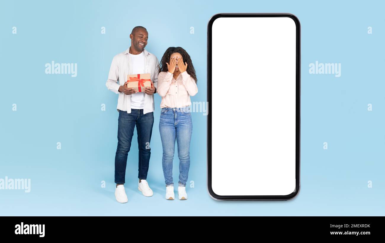 Excited black couple near big smartphone with empty screen, man holding gift box, lady waiting for surprise, mock up Stock Photo