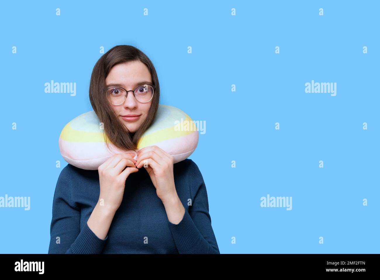Portrait of a student girl in glasses and a sweater sleeping in a travel pillow on a blue background, banner, copy space. A woman at rest with a pillo Stock Photo
