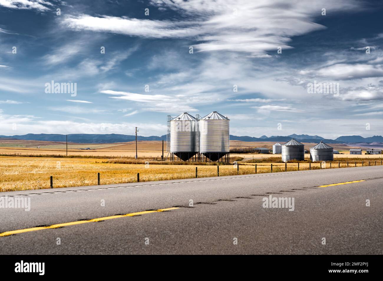 Pair of steel grain silos standing next to a divided highway along harvested fields and distant Rocky Mountains at background near Longview Alberta Ca Stock Photo