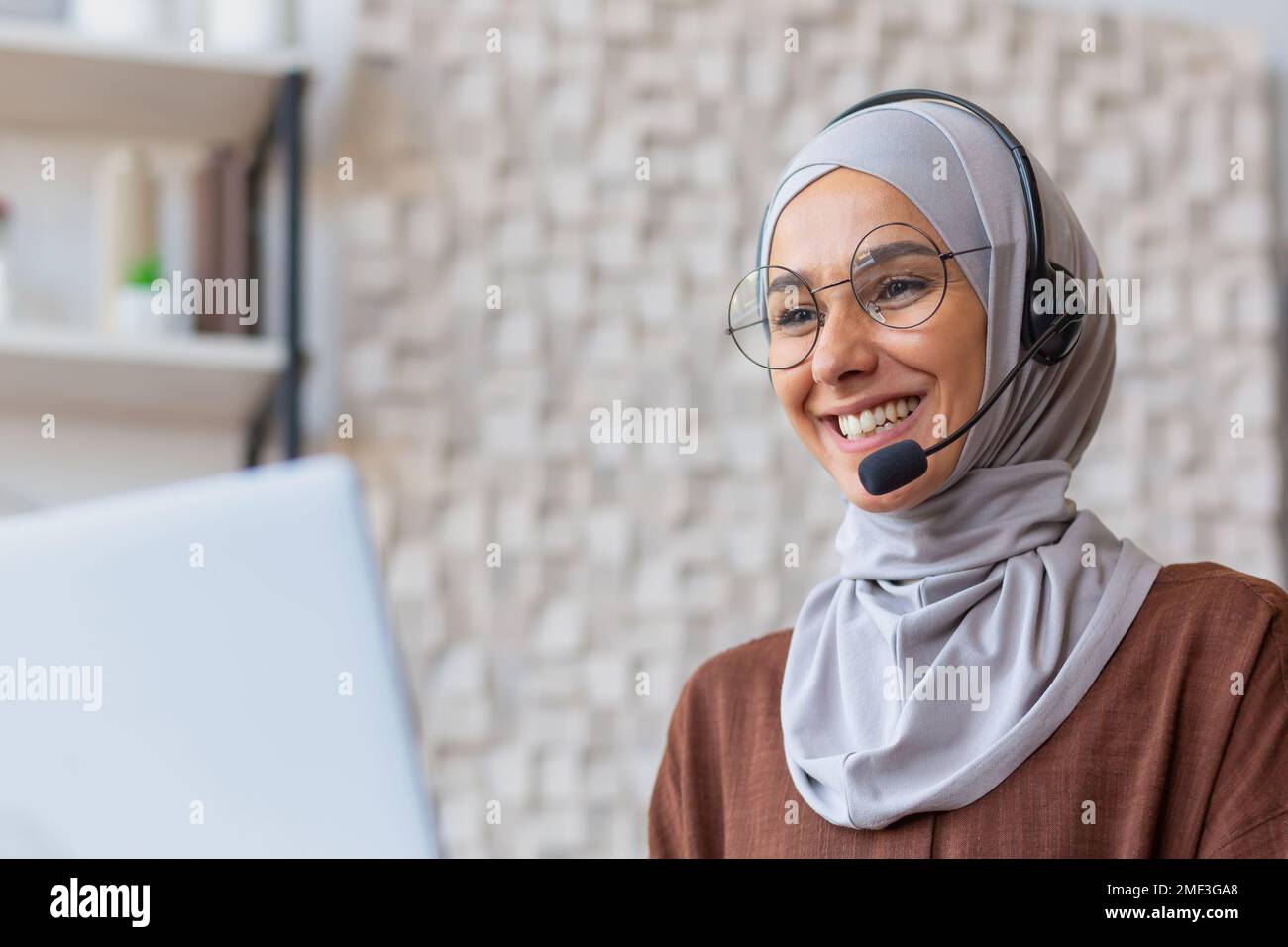 Joyful woman in hijab working from home remotely, Muslim woman talking on video call using headset and laptop, businesswoman working remotely at home. Stock Photo