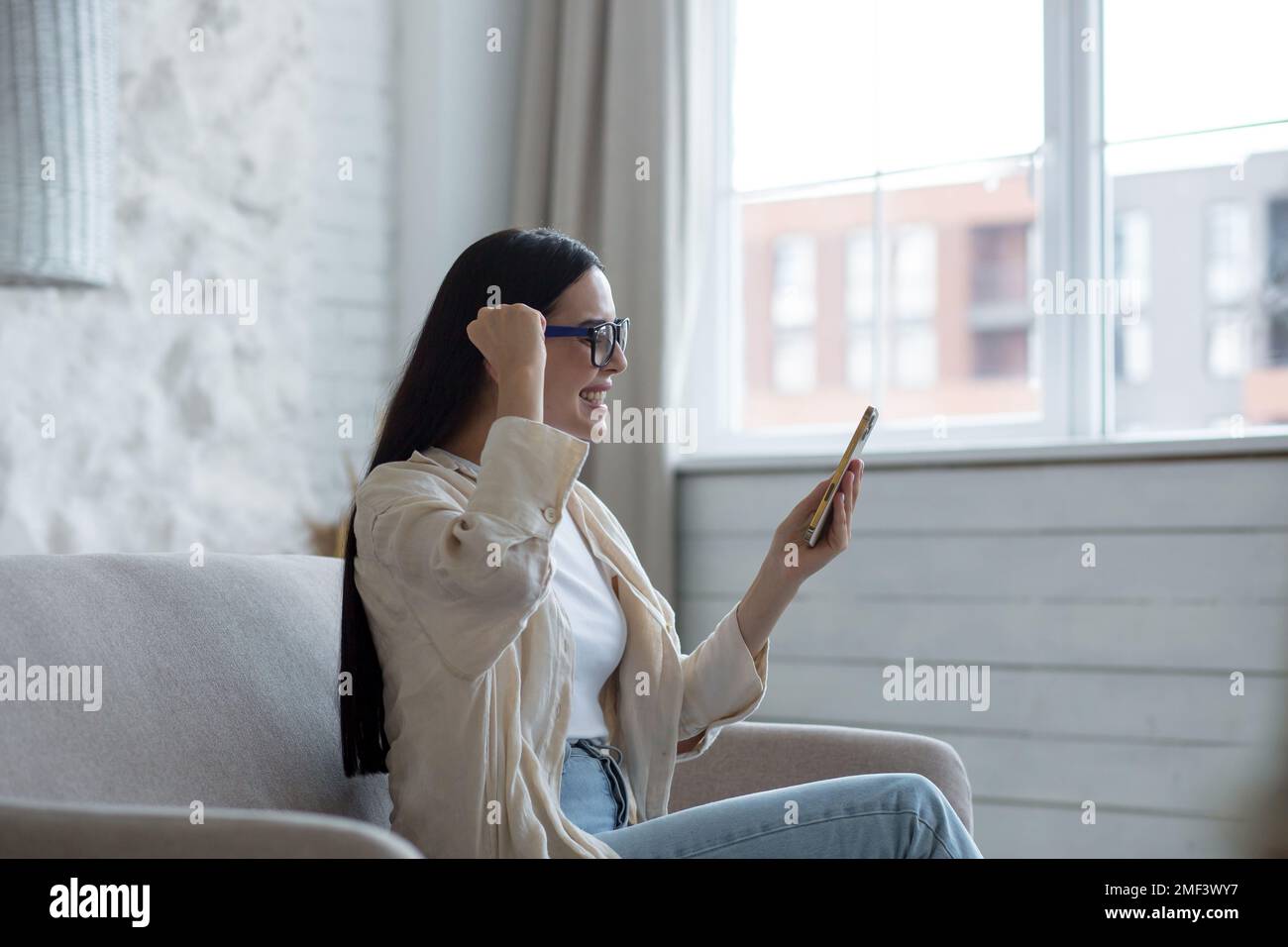 A young woman is sitting at home on the sofa near the window with a phone. plays online games, lottery, raffle. Celebrates the victory, rejoices at the win, shows yes with his hand. Stock Photo