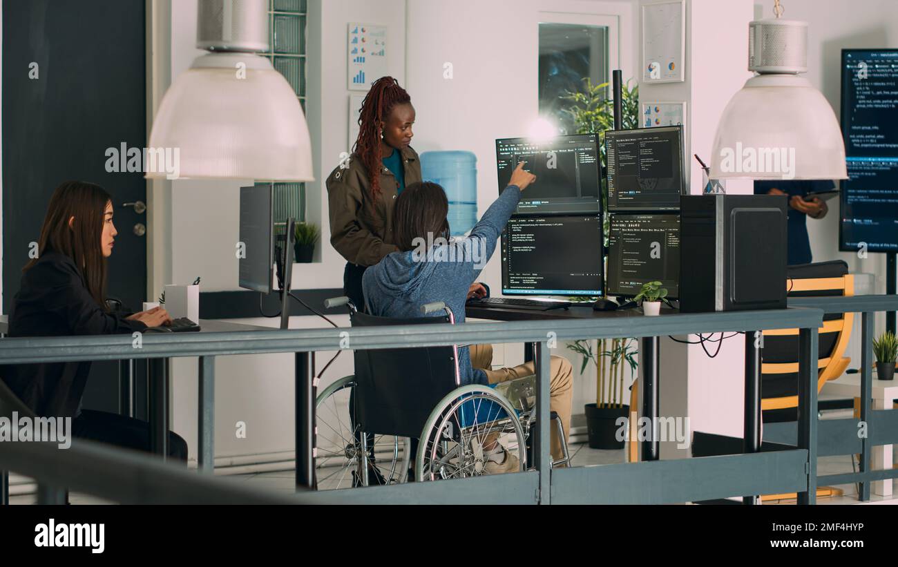 Asian programer with chronic impairment working on software code with it engineer in agency office. App developer with disability in wheelchair using html script on temrinal window. Stock Photo