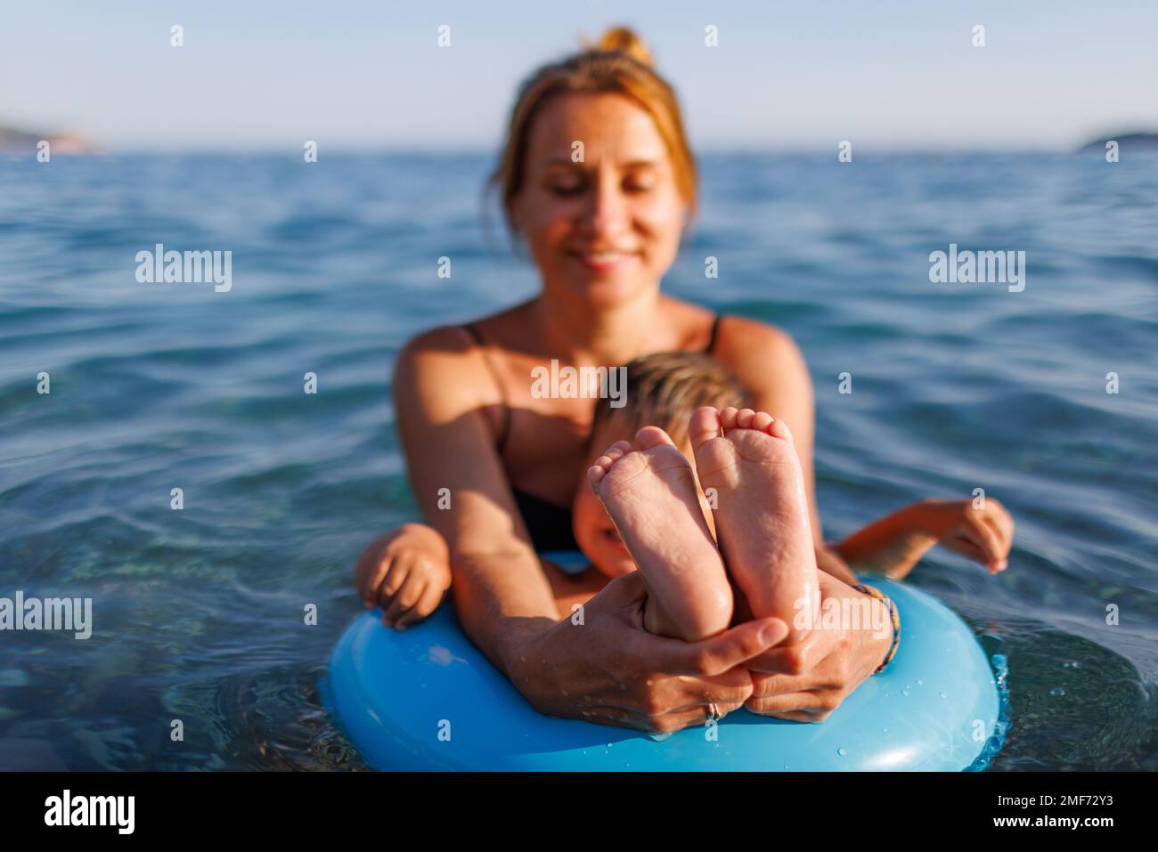 A cute young mother with blond hair smiles and shows to the camera the small tender legs and arms of her baby son, who is sitting in a bright blue inf Stock Photo