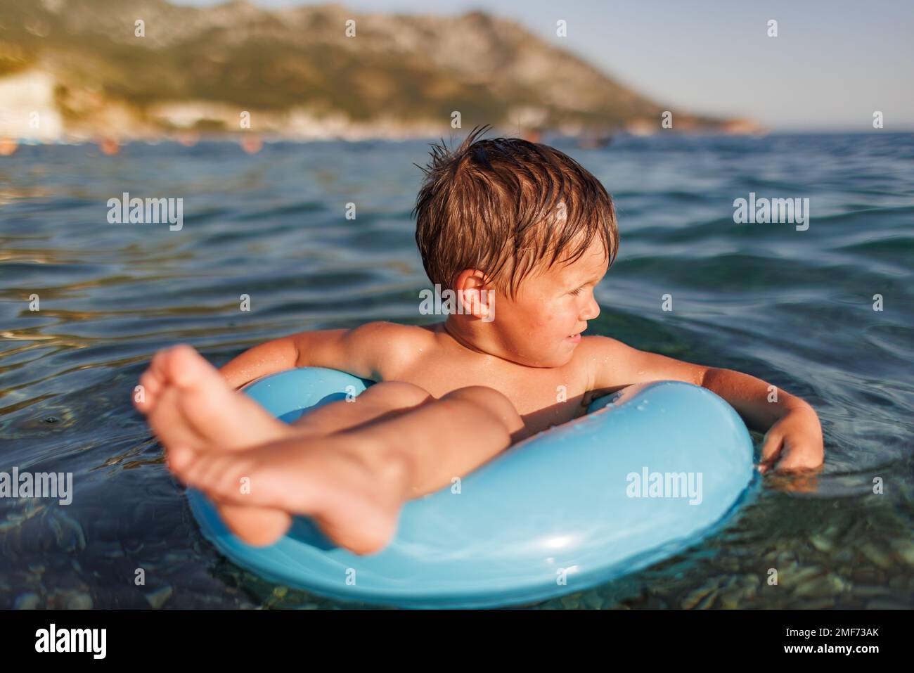 The baby sits in a blue inflatable small circle and looks for his mother, swimming in the blue warm summer Adriatic Sea in the evening sunlight Stock Photo
