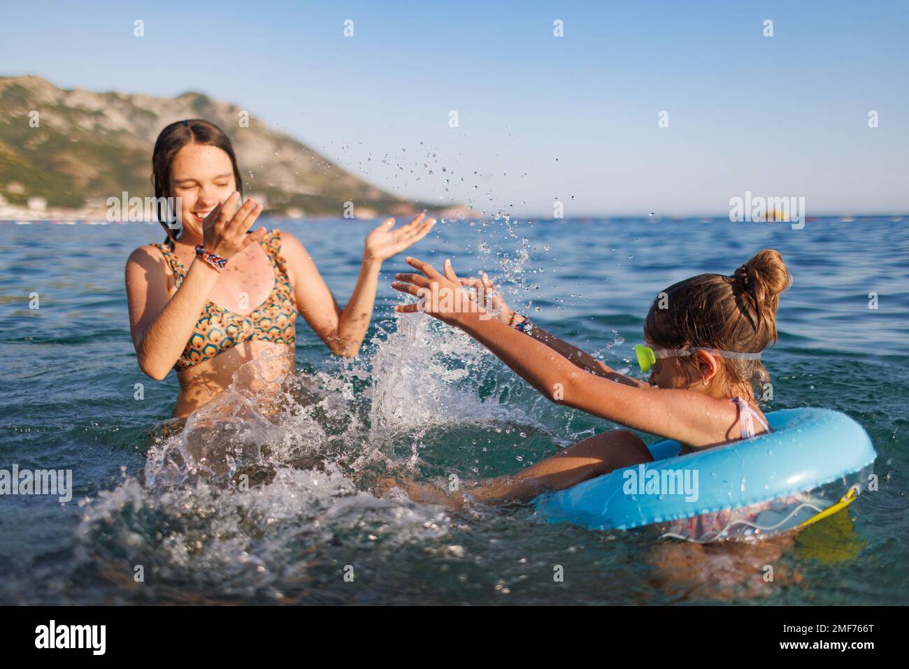 An older sister and her younger sister with blue swimming goggles, who sits in a small bright inflatable circle, splash each other in the warm waters Stock Photo
